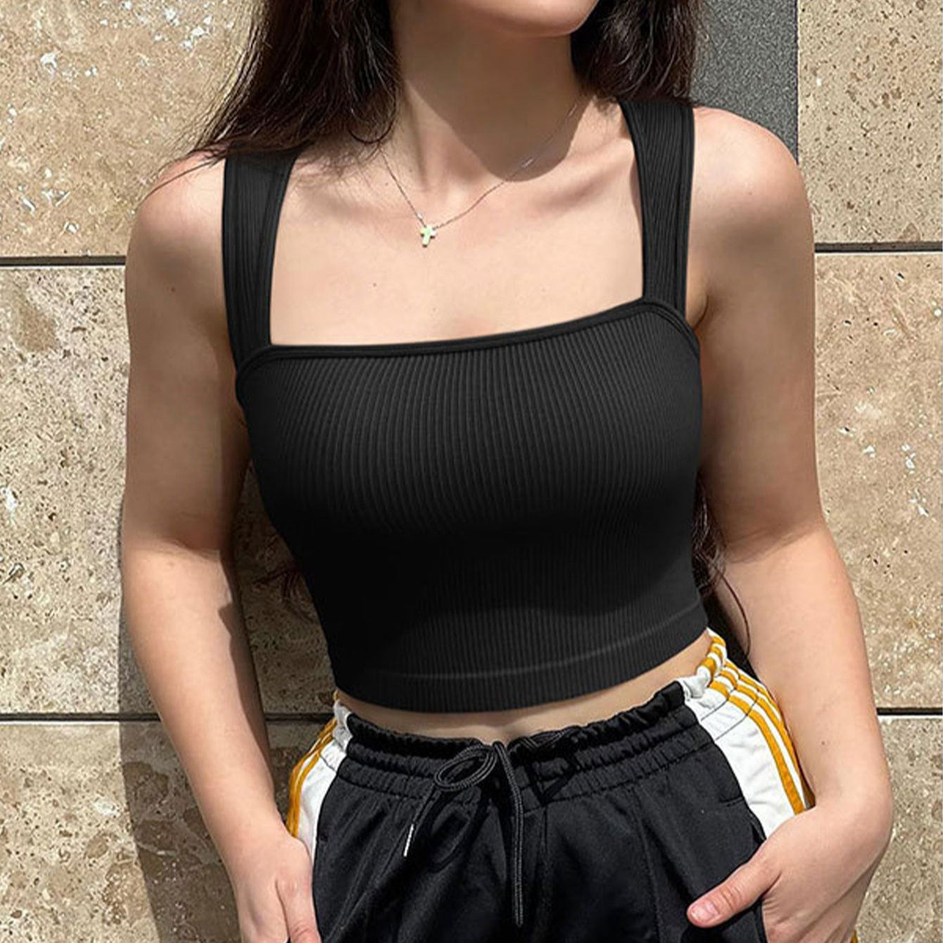 Woman wearing a black crop top and black pants with yellow accents against a stone wall.